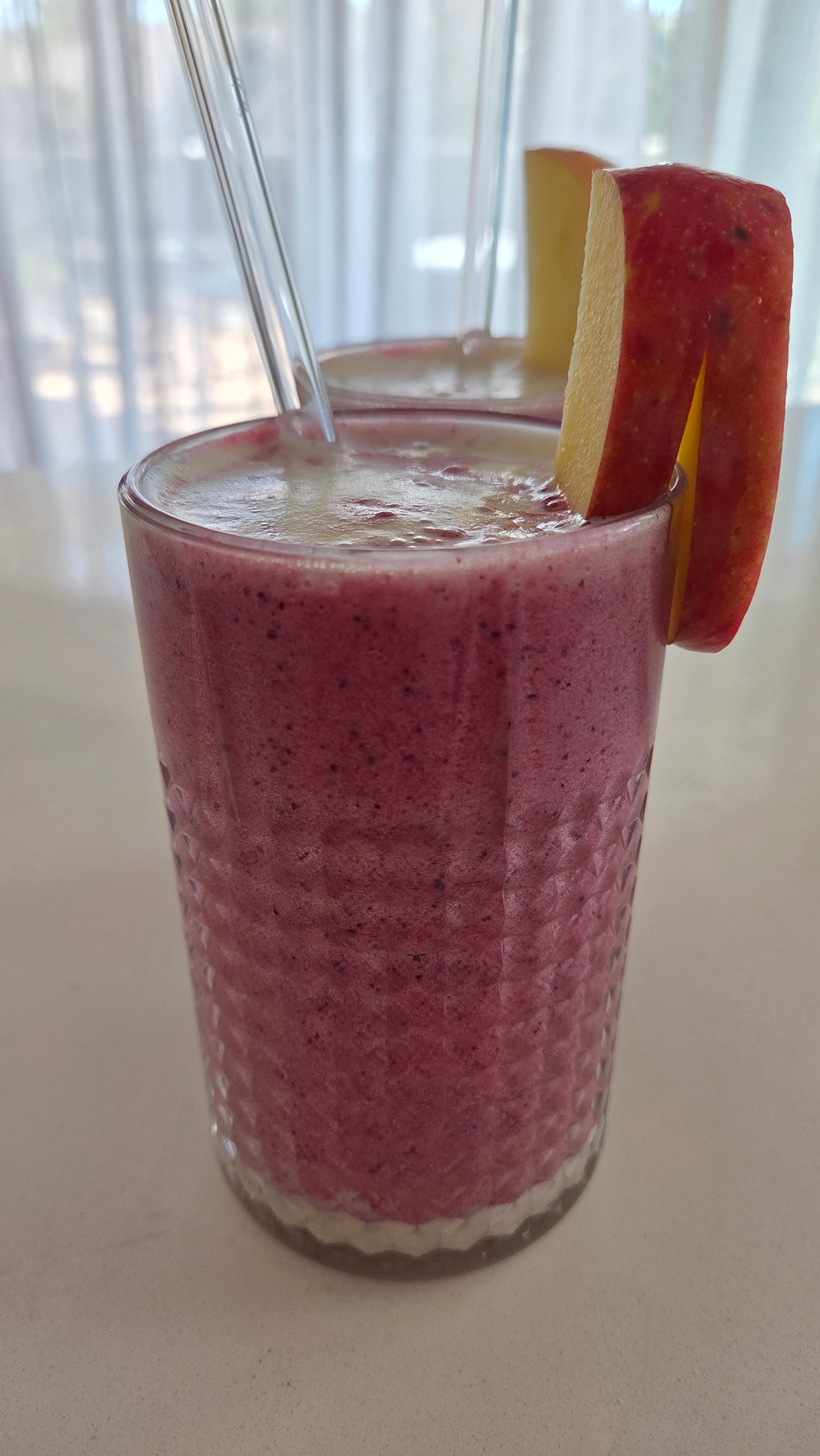 Two tall glasses of the finished Creamy Blueberry Smoothie with oat milk foam and apple slice garnish
