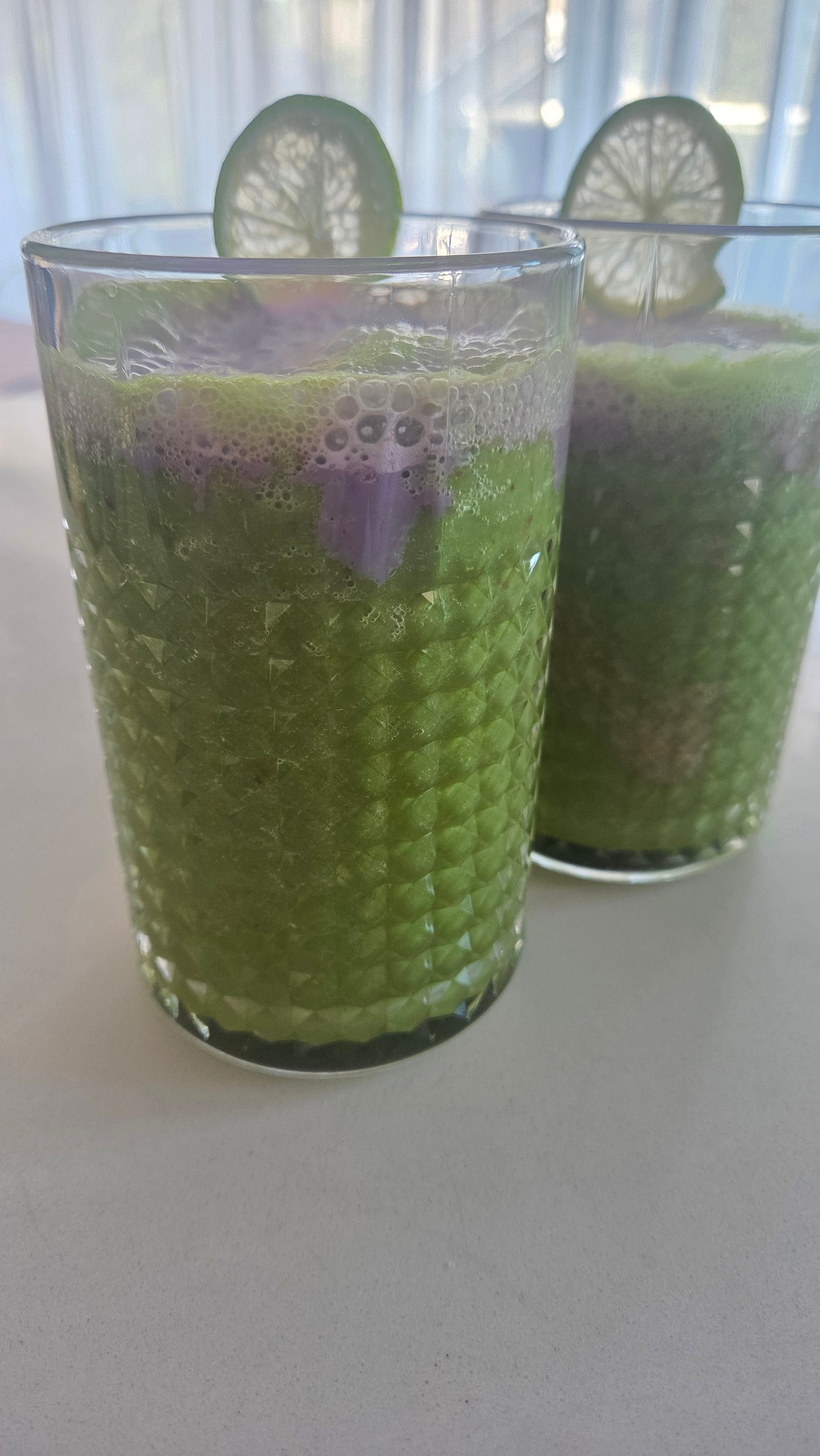 Two diamond-cut glasses of deep green smoothie with purple pitaya foam and lime garnish