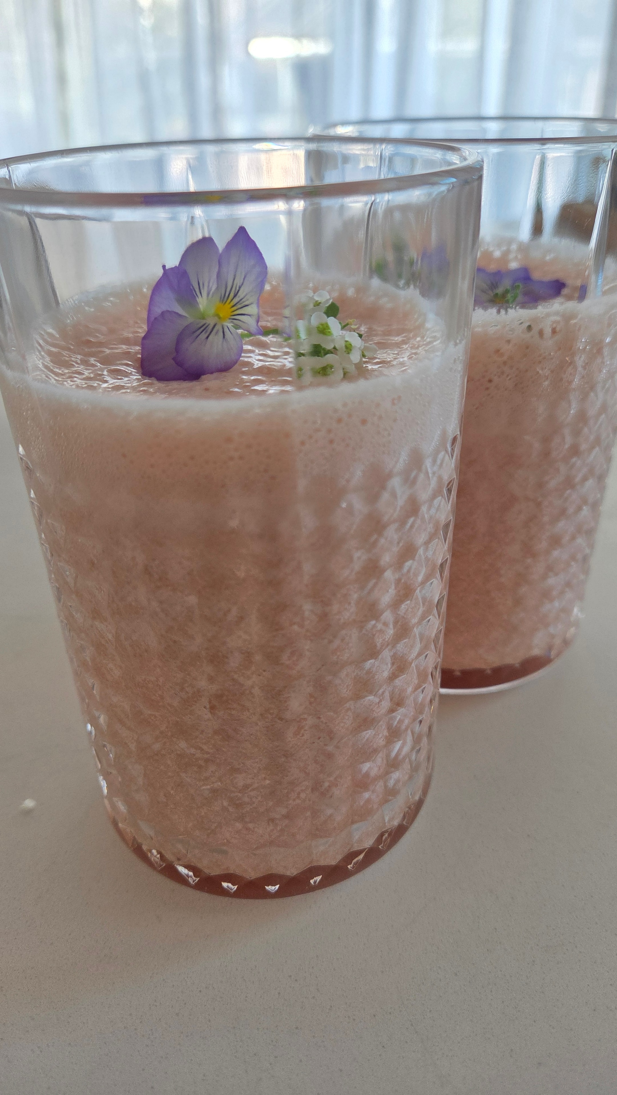 Two glasses of deep rose orange pitaya protein smoothie garnished with viola pansies and white alyssum